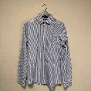 Randig skjorta Tiger of Sweden blå/vit - Klassisk skjorta från Tiger of Sweden i blått och vitt med smala vertikala ränder. Skjortan har lång ärm, knappar framtill och krage. Tillverkad i bomull för en skön känsla och snygg passform. Perfekt för dig som gillar stilrena och tidlösa plagg.