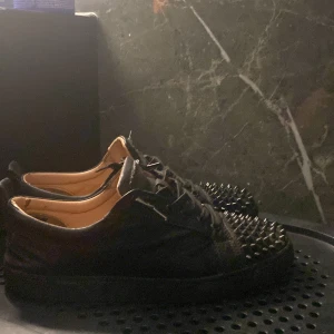 Christian Louboutin svarta sneakers med nitar - Svarta sneakers i skinn från Christian Louboutin med ikoniska röda sulor och coola svarta nitar över tån. Skorna har snörning och en klassisk låg siluett. Perfekt för dig som vill sticka ut med en edgy vibe. Kan sänka priset vid snabbaffär