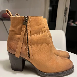 Beiga boots i skinn med klack, stl 39 - Snygga beiga boots i äkta skinn, tillverkade i Spanien. Modellen har en stadig klack, rund tå och dragkedja på sidan med dekorativ rem. Perfekta för dig som gillar en stilren och trendig look med lite höjd.
