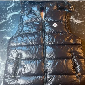 Svart dunväst från Moncler - Snygg svart dunväst från Moncler med glansig finish och klassisk logga på bröstet. Västen har dragkedja framtill, två fickor med dragkedja och hög krage. Perfekt för lager-på-lager och streetstyle. Insidan har ett coolt serietryck.