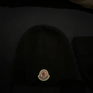Svart Moncler mössa med logga - Svart mössa från Moncler med klassisk rundad form och uppvikt kant. Framtill sitter Monclers ikoniska broderade logga i vitt, rött och blått. Mössan är stickad och har en enkel, stilren look som funkar till alla dina jackor.