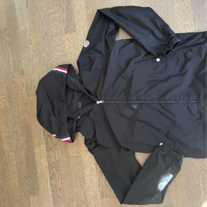 Svart vindjacka från Moncler med huva - Svart vindjacka från Moncler med huva och dragkedja. Jackan har Moncler-logga på ärmen och snygga röd-vita detaljer på huvan. Tillverkad i lätt polyester, perfekt för blåsiga dagar. Klassisk passform och elastiska muddar vid ärmslut.