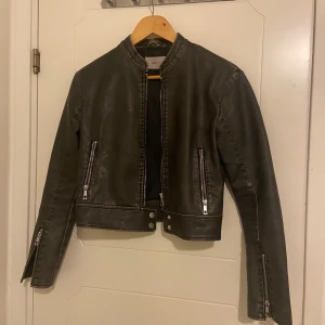 Svart skinnjacka från Nelly - Snygg mörk grön skinnjacka från Nelly med raka linjer och coola dragkedjor på både fickor och ärmslut. Jackan har en kortare, croppad passform. 