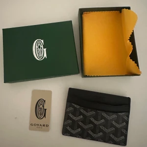 Svart Goyard korthållare i skinn - Exklusiv svart korthållare från Goyard med ikoniskt mönster och diskret logga. Tillverkad i skinn med flera fack för kort. Kommer med originalask och tygpåse. Perfekt accessoar för dig som vill ha stilren och lyxig känsla i vardagen.