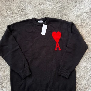 Svart Ami Paris tröja med rött hjärta - Svart stickad tröja från Ami med ett stort rött hjärta och bokstaven A broderat på bröstet. Rund hals och ribbade muddar vid ärmslut och nederkant. Tillverkad i ekologisk bomull, perfekt för dig som gillar stilrena detaljer med en twist.