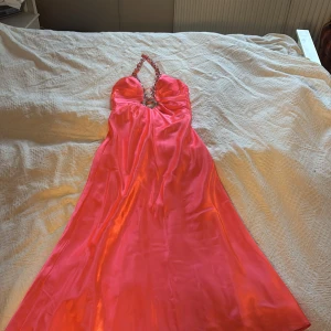 Korallrosa långklänning från Morgan & Co - Klänning perfekt till balen🩷Snygg korallrosa långklänning från Morgan & Co med halterneck och glittriga detaljer längs ringningen. Klänningen har en djup v-ringning med dekorativt smycke framtill och är tillverkad i glansig polyester. Perfekt för dig som vill sticka ut med en färgstark look.