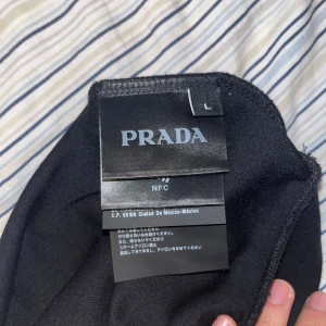 Svart långärmad t-shirt från Prada - Svart långärmad t-shirt från Prada med klassisk rund hals och ikonisk metalltriangel-logga på bröstet. Tillverkad i ett mjukt och glansigt material som känns lyxigt mot huden. Perfekt för dig som vill ha en clean och stilren look med exklusiv känsla.