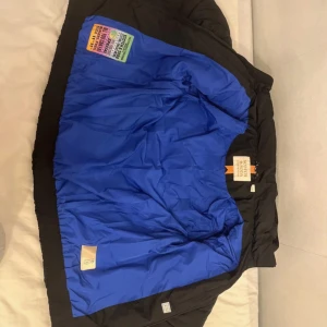 Svart quiltad pufferjacka Scotch & Soda - Svart pufferjacka från Scotch & Soda med cool quiltad design och huva. Insidan är blå och jackan har snygga detaljer som logotyp på ärmen och orange-randig etikett. Perfekt för dig som vill ha en varm och stilren jacka med streetkänsla.