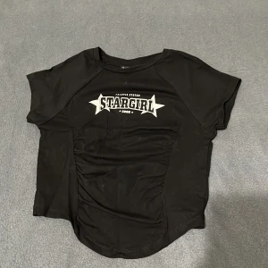 Svart topp med tryck från FB Sister - Svart kortärmad topp från FB Sister med coolt 'STARGIRL' tryck i vitt framtill. Toppen har rynkade detaljer på sidorna och en rund halsringning. Perfekt för dig som gillar streetstyle och vill sticka ut med ett statement-plagg.