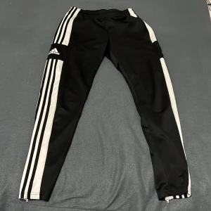 Svarta Adidas träningsbyxor med vita ränder - Adidas träningsbyxor i svart med klassiska vita ränder längs benen och logga på låret. Byxorna har dragkedja vid benslut och elastisk midja. Tillverkade i polyester, perfekt för sport eller chill. Snygg och sportig look med smal passform.