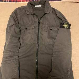 Grå overshirt från Stone Island - Snygg grå overshirt från Stone Island med klassisk krage, två bröstfickor med knappar och ikonisk patch på ärmen. Jackan har hel dragkedja framtill och är tillverkad i bomull. Perfekt för dig som gillar streetwear och vill ha en stilren look.
