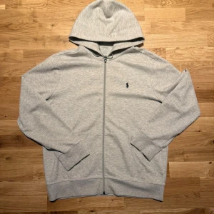Grå hoodie från Polo Ralph Lauren - Snygg grå hoodie från Polo Ralph Lauren med klassisk broderad logga på bröstet. Tröjan har dragkedja framtill, huva och ribbade muddar vid ärmslut och nederkant. Perfekt för chill dagar och enkel att matcha med jeans eller joggers.