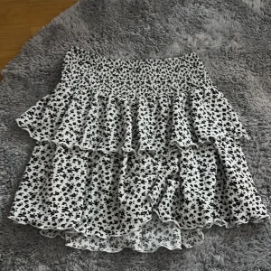 Vit blommig volangkjol från Primark - Supersöt kort kjol från Primark med svart blommigt mönster på vit botten. Kjolen har flera volanger och en smockad midja som ger en skön passform. Perfekt för dig som gillar en feminin och lekfull stil.