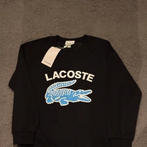 Lacoste tröja - Svart sweatshirt från Lacoste i regular fit med stort vitt Lacoste-tryck och en blå krokodil på bröstet. Tröjan har rund hals, långa ärmar och ribbade muddar vid ärmslut och nederkant. Perfekt för dig som gillar streetwear och tydliga loggor.