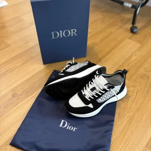 Svarta och vita Dior sneakers - Snygga Dior sneakers i svart och vitt med klassiskt monogrammönster på sidorna. Skorna har vit sula, svarta och vita paneler i mesh och mocka samt vita skosnören. Perfekta för dig som vill ha en lyxig och trendig look.