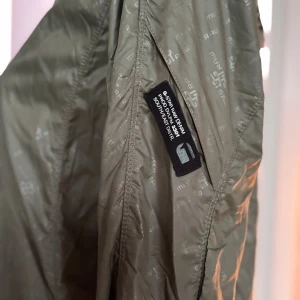 Olivgrön vindjacka från G-Star RAW - Snygg olivgrön vindjacka från G-Star RAW med diskret logomönster över hela jackan. Jackan har hög krage, hel dragkedja framtill och tydlig logga på ryggen. Tillverkad i 100% polyamid, perfekt för blåsiga dagar. Sportig och stilren look.