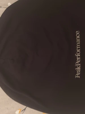 Svart dunväst från Peak Performance - Säljer en svart dunväst från Peak Performance med stilren design och broderad logga på framsidan. Perfekt att slänga på över en hoodie eller tröja när det är lite kyligt ute. Västen har en klassisk passform och är gjord i ett mjukt syntetmaterial.