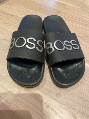 Svarta BOSS sandaler med silvertext - Säljer ett par svarta sandaler från BOSS med bred rem och stor silverfärgad BOSS-logga över foten. Sulan har stjärnmönster undertill och är gjord i syntetmaterial. Perfekta för chill dagar och enkel att dra på sig.