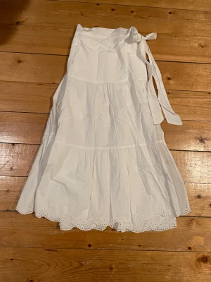 Söt långkjol från Lindex✨ - Säljer en vit långkjol från Lindex i storlek XS. Kjolen har knyt i midjan, volanger och broderade spetsdetaljer längst ner. Tillverkad i bomull, perfekt för ljusa dagar. Kjolen har en omlott-design och är riktigt flowy och skön✨☺️