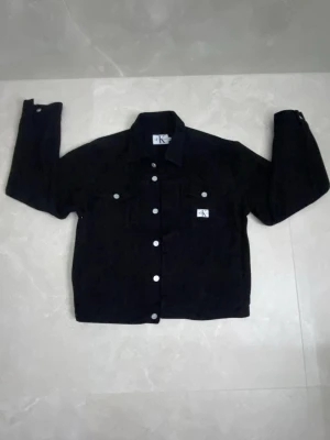 Svart jeansjacka från Calvin Klein - Svart jeansjacka från Calvin Klein Jeans i klassisk modell med silvriga knappar framtill och på ärmsluten. Jackan har två bröstfickor med lock och CK-logga på vänster sida. Perfekt för dig som gillar stilrena och tidlösa plagg.