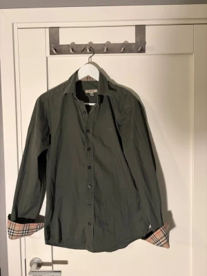 Grön Burberry skjorta med rutiga detaljer - Snygg grön skjorta från Burberry i bomull med klassiska rutiga detaljer på insidan av krage och manschetter. Skjortan har långa ärmar, knappar framtill och broderad logga på bröstet. Perfekt för dig som gillar stilrena plagg med lyxig känsla.