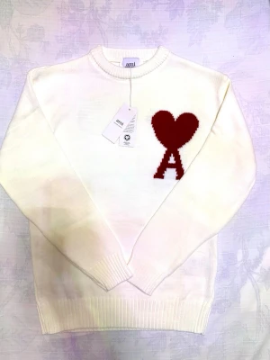 Ami Tröja  - Hej! Säljer en vit stickad tröja från Ami som har den ikonsika röda hjärtat och bokstaven A på bröstet. Tröjan har rund hals, ribbade muddar och lång ärm. Perfekt för dig som gillar stilrena detaljer och vill ha något som sticker ut lite extra. Passar perfekt till vardagen men även till festliga tillfällen 😊❄️
