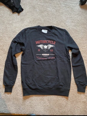 Svart sweatshirt från DNMD med tryck - Cool svart sweatshirt från DNMD med tryck framtill som har motorcykeltema och texten 'Motorcycle Legendary Riders'. Tröjan har rund hals och långa ärmar, perfekt för en avslappnad stil.