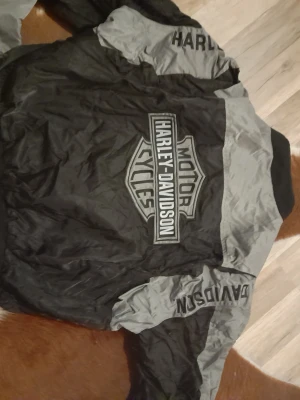 Harley-Davidson svart/grå vindjacka 2XL - Säljer en svart och grå vindjacka från Harley-Davidson i storlek 2XL. Jackan har stora loggor på ryggen och ärmarna, dragkedja framtill och coola detaljer. Tillverkad i polyester, perfekt för dig som vill sticka ut på hojen.