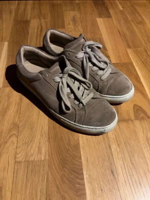 Beigea mockasneakers från Les Deux - Snygga beige bruna sneakers från Les Deux i mjuk mocka med vita sulor och snörning. På hälen finns broderade detaljer i blått, vitt och rött. Skorna har en klassisk, rund tå och platt sula som passar till många olika stilar.
