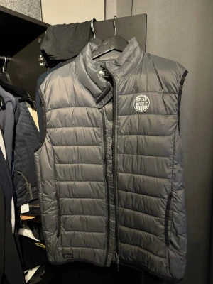 Grå dunväst från Rögle - Säljer en grå dunväst från Rögle med quiltad design och dragkedja framtill. Västen har hög krage och två fickor på sidorna. Perfekt för lager-på-lager och sportig stil. Rögle-logga på bröstet ger en snygg detalj.