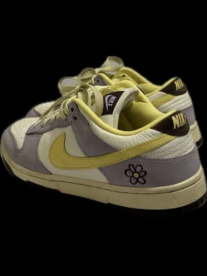 Nike Dunk Low Pastellila & Gul Blomma - Nike Dunk Low sneakers i pastellila, gul och vitt med broderad blomma på hälen. Skorna har klassisk snörning, perforerad tåbox och svart yttersula. Ovandelen är i mocka och skinn för en schysst mix av material och färger. Perfekt för dig som vill sticka ut med en unik sneaker.