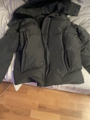 Svart pufferjacka från Canada Goose L - Säljer en svart pufferjacka från Canada Goose i storlek L. Jackan har huva, dragkedja och knappar framtill samt två stora fickor. Perfekt för kalla dagar och riktigt skön att bära. Klassisk design med diskret logga på insidan.