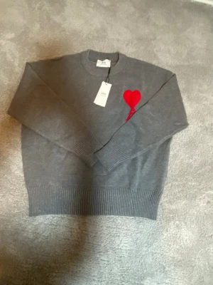 Grå tröja med rött hjärta från Ami - Säljer en grå tröja från Ami Alexandre Mattiussi med ett ikoniskt rött hjärta broderat på bröstet. Tröjan har rund hals, ribbade muddar och är tillverkad i mjuk bomull. Perfekt för dig som gillar stilrena detaljer och vill ha något som sticker ut.