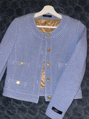 Blå rutig kavaj från Morris Lady - Snygg blå kavaj från Morris Lady med vitt rutmönster och guldiga knappar. Jackan har rund halsringning, fyra fickor framtill och är helfodrad med beige insida. Perfekt för dig som vill ha en klassisk men trendig look.