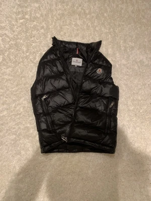 Svart dunväst från Moncler - Säljer en svart dunväst från Moncler med glansig finish och klassisk quiltad design. Västen har två dragkedjeförsedda fickor framtill och en hög krage. Perfekt för lager-på-lager och snygg till både hoodie och t-shirt.