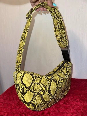 Gul axelväska från Beck Söndergaard - Säljer en riktigt cool axelväska från Beck Söndergaard i gul och svart ormmönstrad design. Väskan har justerbar rem och dragkedja upptill. Materialet är syntet och väskan har en mjuk, formad siluett som sticker ut i mängden.
