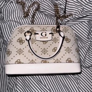 Beige handväska från Guess med kedja - Säljer en beige handväska från Guess med guldfärgad kedjerem och klassiskt logomönster i brunt. Väskan har två handtag, dragkedja upptill och en praktisk ficka på baksidan. Insidan är fodrad med Guess-tryck och har flera fack för småsaker.