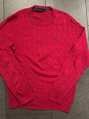 Röd kabelstickad tröja Morris XL - Kabelstickad röd tröja från Morris i storlek XL. Klassisk rund halsringning och diskret logga broderad på bröstet. Tröjan har långa ärmar och ribbade muddar. Perfekt till höst och vinter för en stilren look.