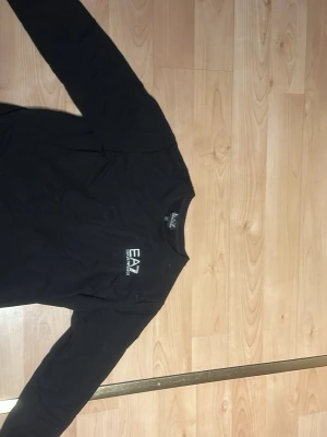 Svart långärmad tröja EA7 Armani - Svart långärmad tröja från EA7 Emporio Armani med rund hals och vit logga på bröstet. Tröjan är i mjuk bomull och har en clean, stilren look som passar perfekt till jeans eller joggers.