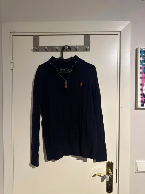 Marinblå stickad polotröja Polo Ralph Lauren - Säljer en marinblå stickad polotröja från Polo Ralph Lauren med klassiskt kabelstickat mönster och röd broderad logga på bröstet. Tröjan har half zip-dragkedja och ribbad krage, perfekt för kyliga dagar.