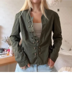 Olivgrön utilityjacka med knappar - Snygg olivgrön utilityjacka med coola knappar framtill och justerbara spännen i midjan. Jackan har lång ärm och en rak passform som funkar till många olika outfits. Perfekt för dig som gillar en avslappnad men ändå stilren look.