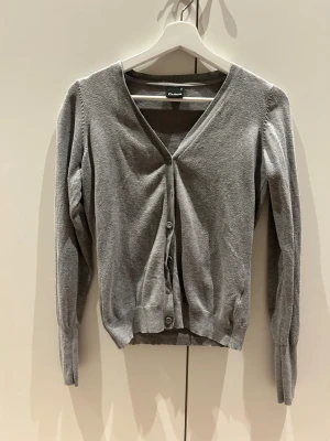 Grå kofta med knappar - Säljer en stilren grå kofta från Cubus i mjuk bomull. Koftan har v-ringning, långa ärmar och knappar framtill samt extra knappar vid ärmsluten för en snygg detalj. Perfekt att slänga över en topp under kyligare dagar.
