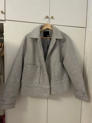 Grå ulljacka från 157 - Snygg grå jacka i ullblandning från 157 med klassisk krage, stora framfickor och knäppning med knappar. Jackan har en boxig, lite loose passform och är helfodrad. Perfekt för dig som gillar stilrena och enkla plagg med en modern touch.