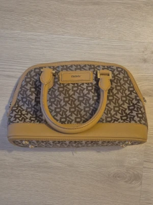 Beige och brun DKNY handväska - Säljer en snygg ÄKTA handväska från DKNY med klassiskt logomönster i brunt och beige. Väskan har två handtag, guldfärgade detaljer och dragkedja upptill. Utsidan är i canvas med skinnliknande detaljer och insidan har fack för småsaker.