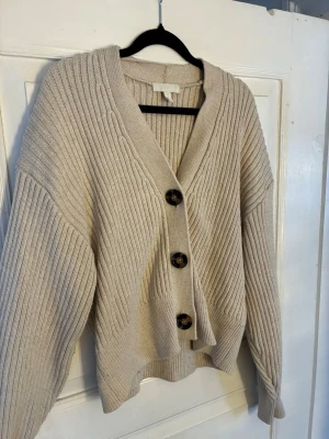Beige ribbstickad kofta från H&M - Supermysig beige kofta från H&M i ribbstickat material. Koftan har stora svarta knappar framtill, v-ringad hals och en loose fit. Perfekt att slänga på sig när det är kyligt ute. Passar XS/S