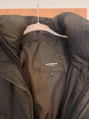 Svart lång pufferjacka Rockandblue - Säljer en svart lång pufferjacka från Rockandblue med hög krage och dragkedja samt knappar framtill. Jackan har två fickor med dragkedja och Rockandblue-logga på ärmen. Perfekt för kalla dagar och riktigt snygg till vintern.
