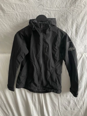 Svart vindjacka från McKinley - Säljer en svart vindjacka från McKinley med huva och dragsko. Jackan har hel dragkedja framtill, en bröstficka med dragkedja och meshfoder på insidan. Märkeslogga på ärmen och ryggen. Perfekt för utomhusaktiviteter när det blåser.