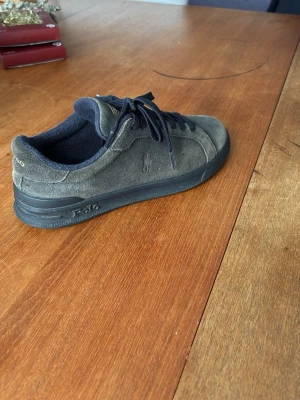 Grå mocka sneakers från Polo Ralph Lauren - Säljer ett par snygga sneakers från Polo Ralph Lauren i grå/blå mocka med mörkblå detaljer och snörning. Skorna har en klassisk låg profil, svart sula och diskret Polo-logga på sidan och sulan. Perfekta till jeans eller chinos för en clean look.