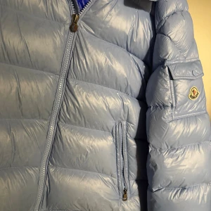 Ljusblå Moncler Maya - Säljer en ljusblå Maya från Moncler med ikonisk logga på ärmen och quiltad design. Jackan har dragkedja framtill, ficka med lock på ärmen och innerfoder i stark blå färg med serietidningsdetalj. Perfekt för kalla dagar och riktigt snygg streetstil. Storlek 5 vilket passar XL bäst. OBS! Lapparna är klippta. Jackan skulle behöva en kemtvätt, annars väldigt bra skick. Returer accepteras inte!!