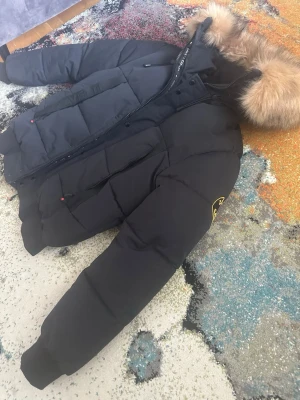 Svart pufferjacka från Kanad - Säljer en svart pufferjacka från Kanada Goose med stor fluffig päls runt huvan. Jackan har dragkedja, ribbade muddar och två stora fickor framtill. Klassisk patch på ärmen och riktigt varm design, perfekt för kalla dagar.